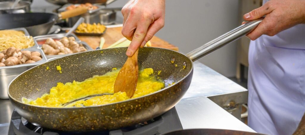 Primo piano di uno chef che cuoce uova strapazzate in una padella sul fuoco con una spatola di legno