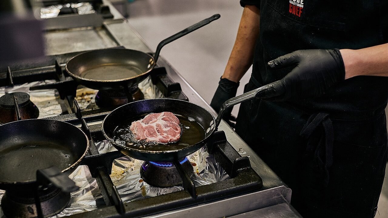 Chef con guanti neri impugna una padella in ferro lionese sul fuoco mentre cuoce una bistecca