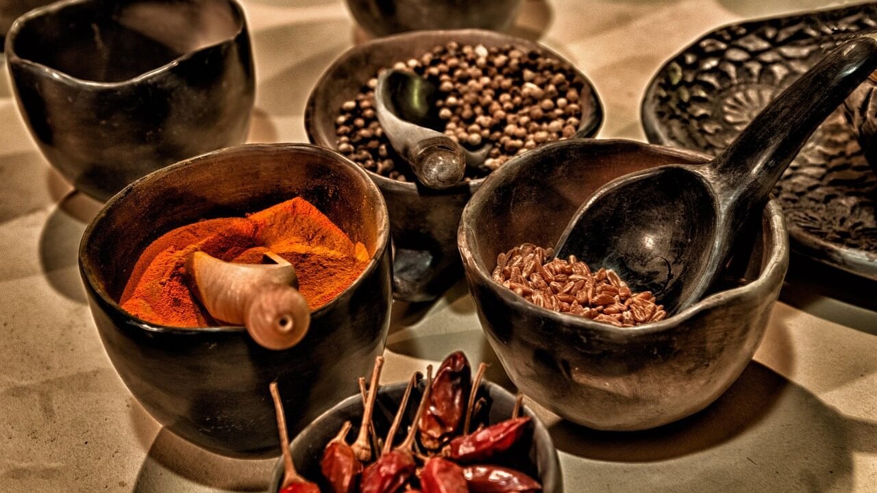 Composizione di ciotole in ceramica scura contenenti spezie varie, peperoncini e semi con cucchiai in legno, che rappresenta la ricerca dell'identità del gusto e dell'intensità naturale